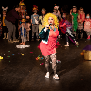 a drag performer on stage talking into a microphone with children standing in the background
