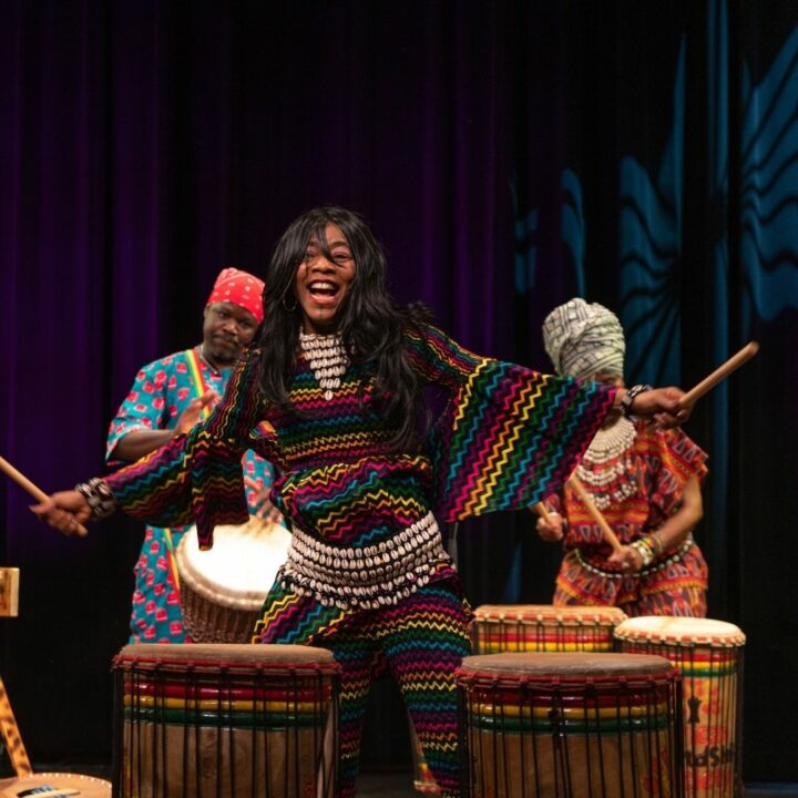 Performer from Africa Oyé performance, wearing a colourful outfit and smiling holding drumsticks in the air