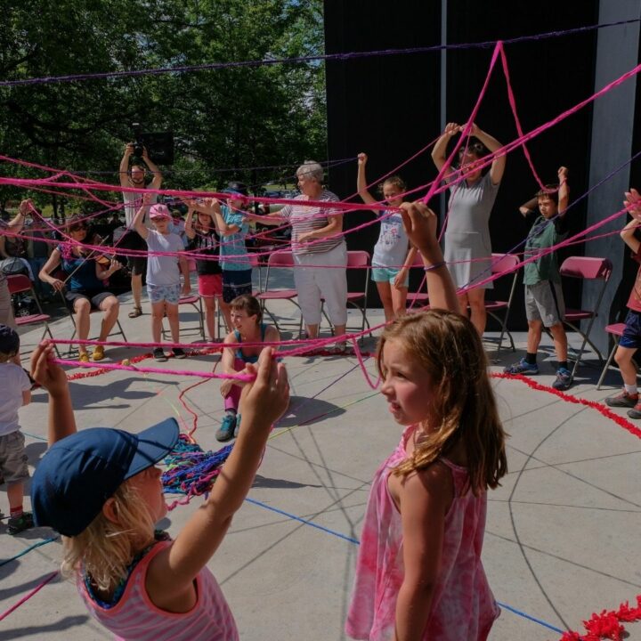 Image of a group of children holding pink yarn in the air during our Tricoter dance performance on May 31st through to June 5th