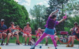 Image of woman in purple leaning to the side with her arms in the air, holding a pink ball of yarn. Join us for our Tricoter performance May 31st-June 5th