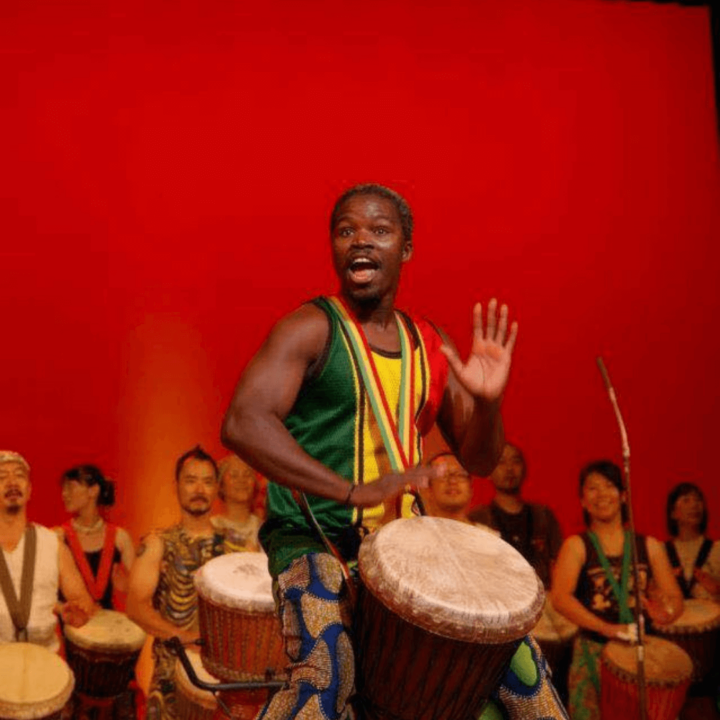 Image of performer from the Africa Oyé performance singing and drumming in front of a red background