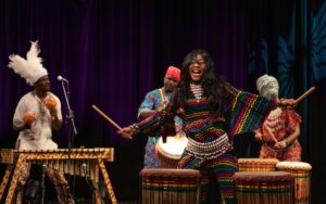 Performer from Africa Oyé performance, wearing a colourful outfit and smiling holding drumsticks in the air