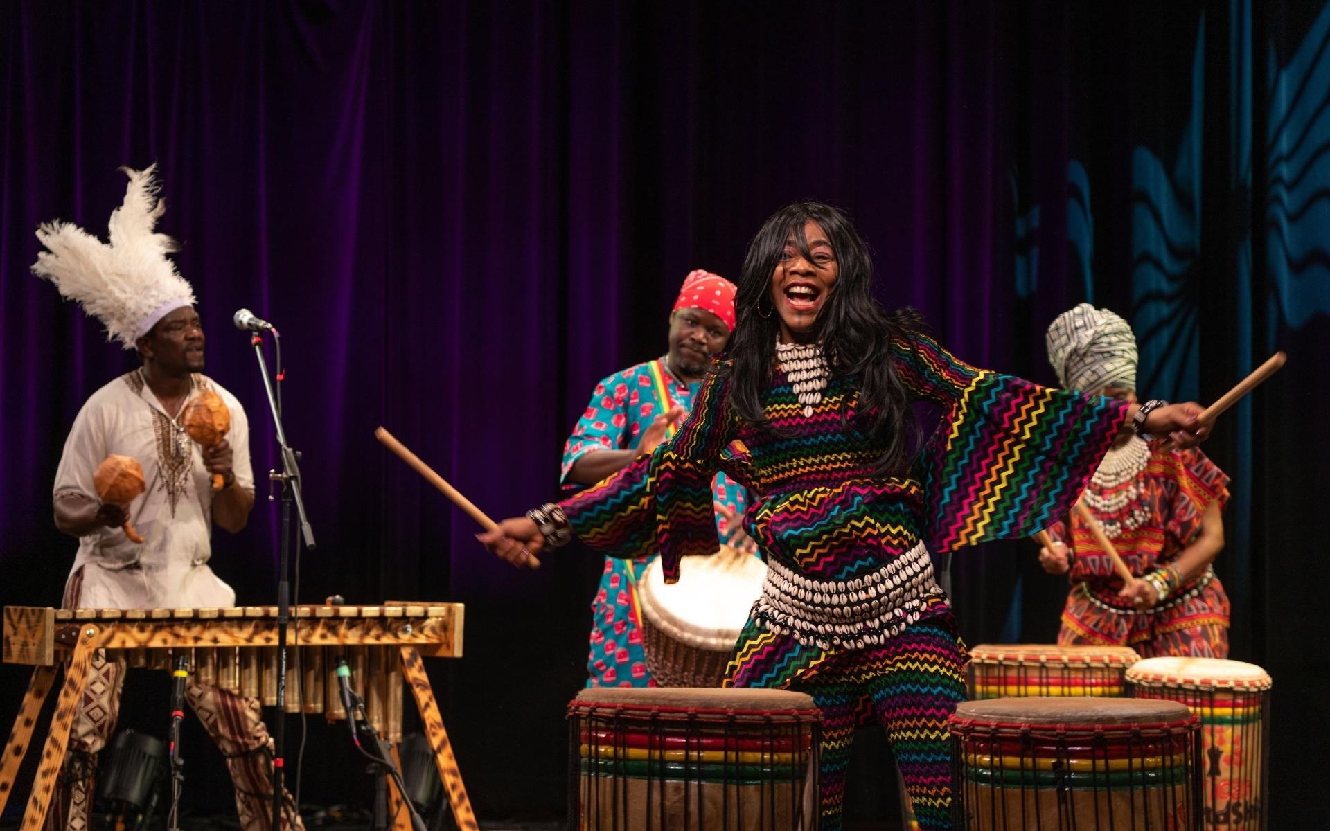 Performer from Africa Oyé performance, wearing a colourful outfit and smiling holding drumsticks in the air