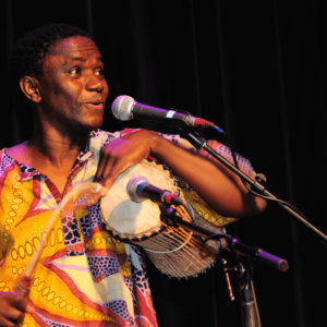 Image of a performer from the Africa Oyé performance wearing yellow, singing and drumming into microphones