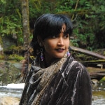 Image of Anjalica Solomon standing in the forest. They are a featured artist in the future artists performance on June 3rd