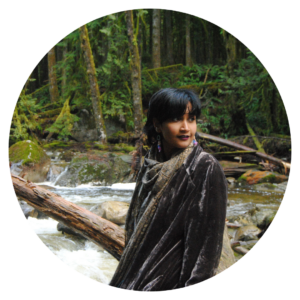 Image of Anjalica Solomon standing in the forest. They are a featured artist in the future artists performance on June 3rd