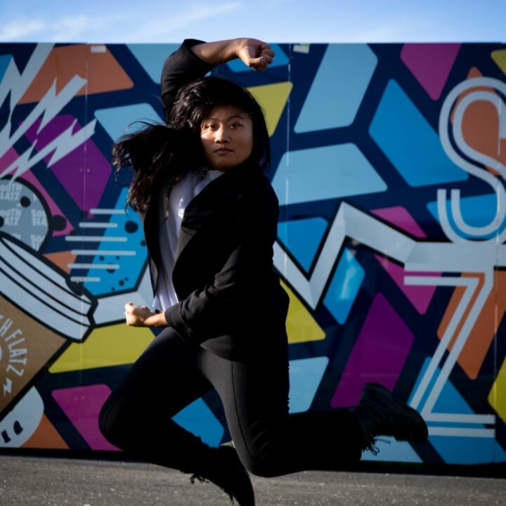 Image of Find Mutya jumping in the air in front of street art. She is a featured artist in the future artists performance on June 3rd