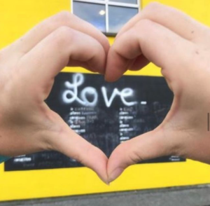 Photo of a Mural of the word Love in white letters with a black background. Someone has put their hands in the shape of a heart to surround the text in the photo