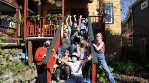 2021 cast of One of a Kind: sitting together on back stairs of house in the sunshine. Wearing masks with their hands in the air