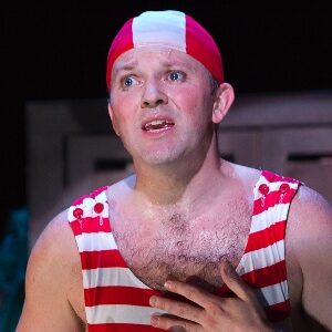 Actor Louis Lovett stands in the spotlight with a red and white striped old fashioned swim suit and cap, hands on heart, and mouth open