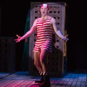 Actor Louis Lovett stands in the spotlight with a red and white striped old fashioned swim suit arms outstretched and mouth open wide in song