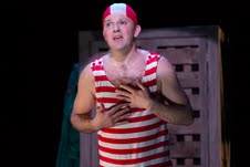 Actor Louis Lovett stands in the spotlight with a red and white striped old fashioned swim suit and cap, hands on heart, and mouth open