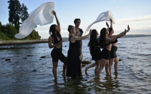 Image of the performers from the Living With performance wearing black and standing in shallow water with their hands up to the sky. Two members holding flowy white cloth and a fan