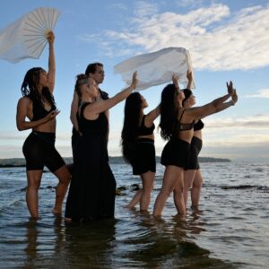 Image of the performers from the Living With performance wearing black and standing in shallow water with their hands up to the sky. Two members holding flowy white cloth and a fan
