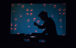 Image of the outline of a man looking down in front of a screen with red shapes on it for the ‘A Story of a House that Turned into a Dot’ performance