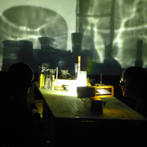 Image of a desk with objects and shadows on the wall for the ‘A Story of a House that Turned into a Dot’ performance