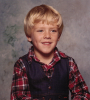 A childhood photo of one of our board members, Jason Dubois, smiling in front of a gray backdrop, wearing a red plaid shirt and a black vest.