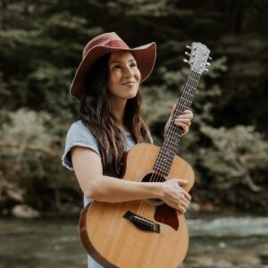 Musician Gina Lina in natural setting holding guitar and smiling to someone off camera. Performing on Saturday June 4.