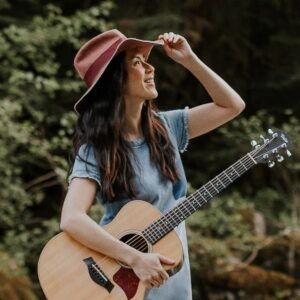 Ginalina outside in natural setting, holding guitar with one hand and grabbing the edge of her hat with the other as she looks up and out. Performing on Saturday June 4.