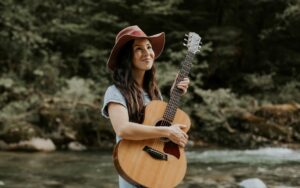 Image of Ginalina wearing a red hat smiling and looking up while holding a guitar in front of a river in the forest