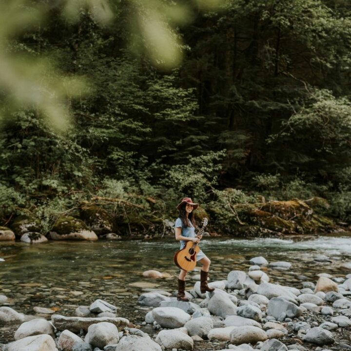 Image of Ginalina holding a guitar standing beside a river in the forest