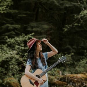 Image of Ginalina holding the brim of her hat and looking up while holding her guitar in the forest
