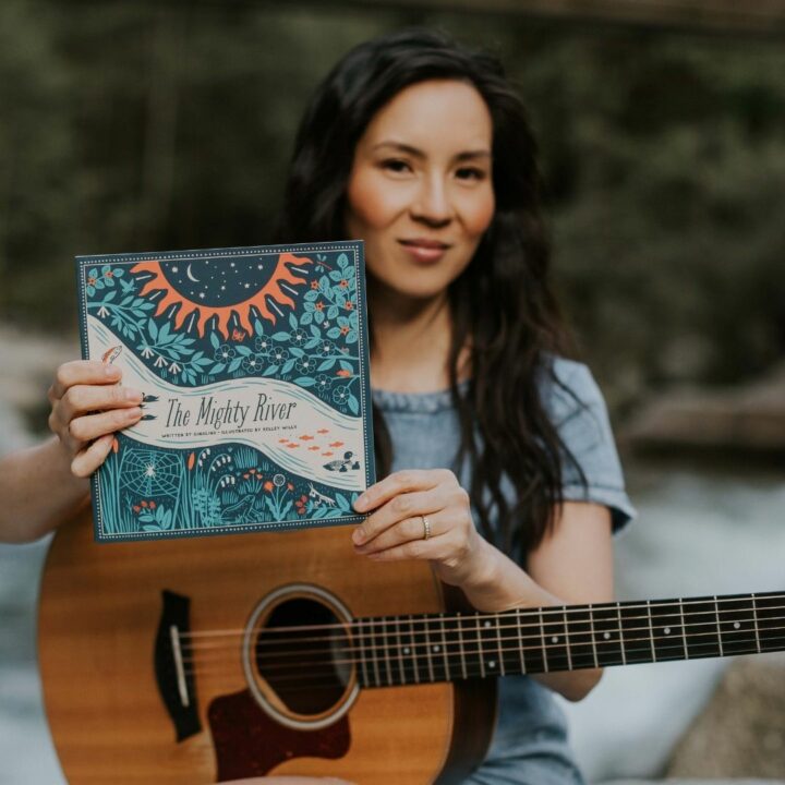 Image of Ginalina holding a guitar and holding up her book, The Mighty River in front of a river in the forest