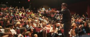 Image of Fred Penner waving at an audience in red auditorium chairs