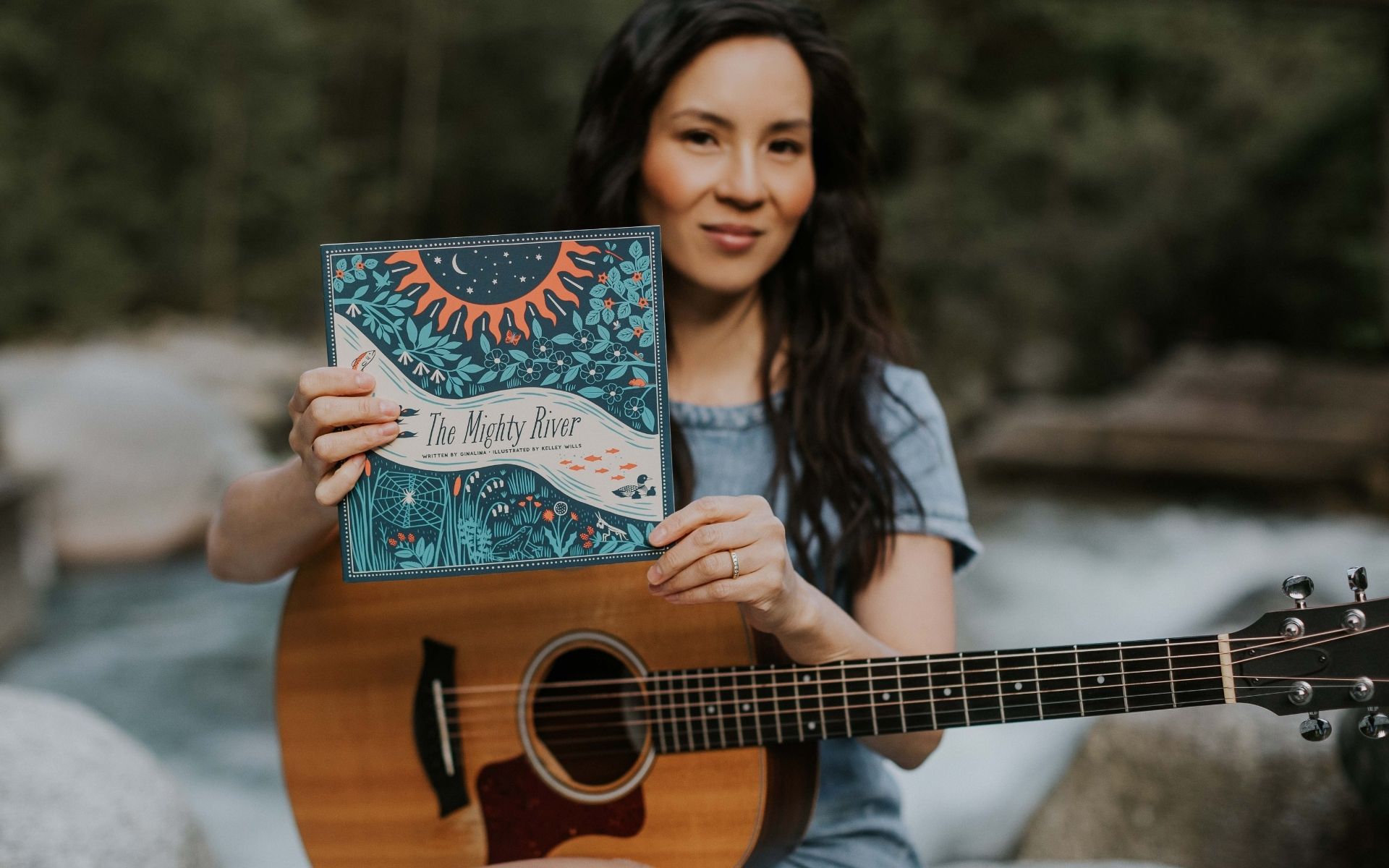 Ginalina sitting beside a river with her guitar, holding up her book The Mighty River. Join us for Ginalina’s workshop, The Wild & Wonderful West Coast workshop on June 5th