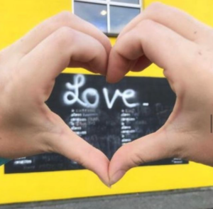 Two hands held in the shape of a heart in front of a yellow wall with ‘love’ written on it on a chalk board