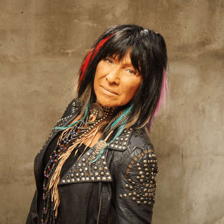 Buffy Sainte Marie, wearing a black leather jacket, standing in front of a brown backdrop
