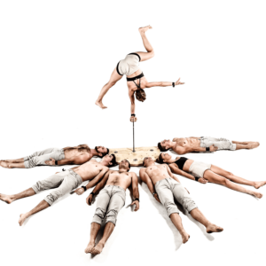 6 performers lying on the floor, while 1 performer is balancing upside down on a thin pole