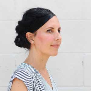 Image of a women smiling, wearing a striped shirt,in front of a white brick wall