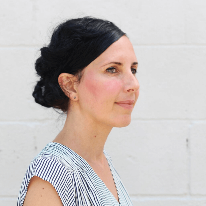Image of a women smiling, wearing a striped shirt,in front of a white brick wall