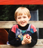 A childhood photo of one of our board members, Cameron Wilson, smiling while holding a partially eaten apple in their hands.