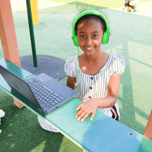 a child smiling while sitting in front of a laptop and wearing a white shirt and green headphones