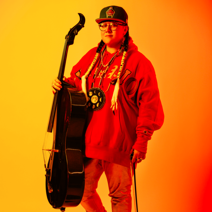 Cris Derken holding a cello, while standing in front of an orange backdrop