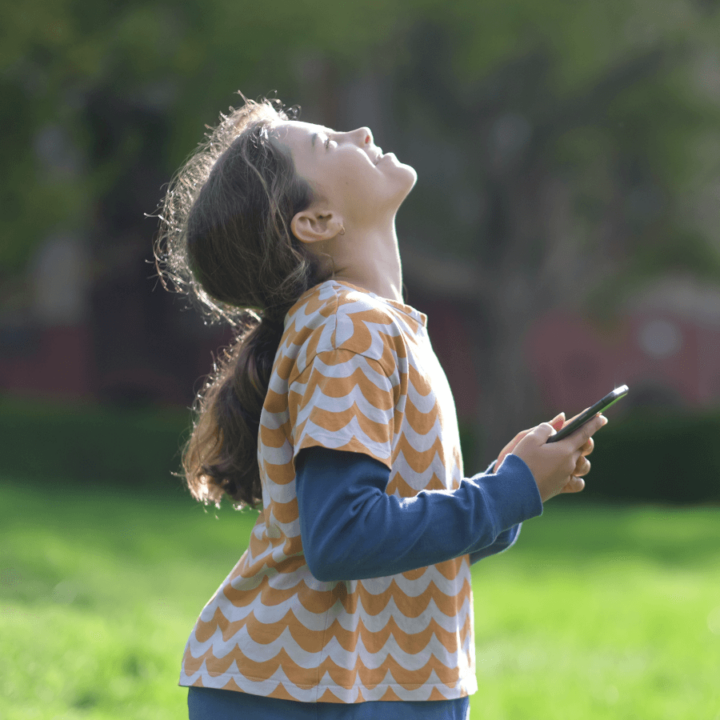 girl outside looking up at the sky with sun shining on her face while holding device.