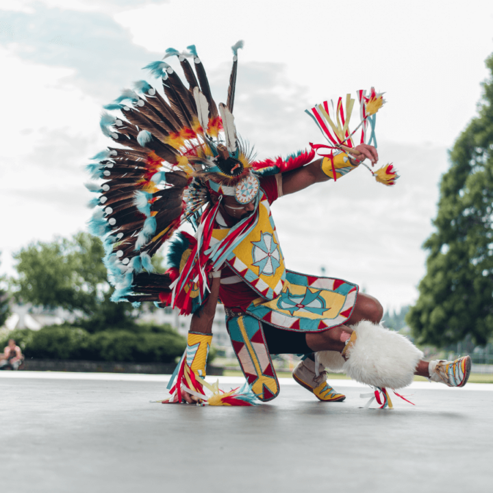 one of the dancers from the dance group The Wild Moccasin Dancers, dancing on stage and wearing Indigenous regalia, with trees in the background