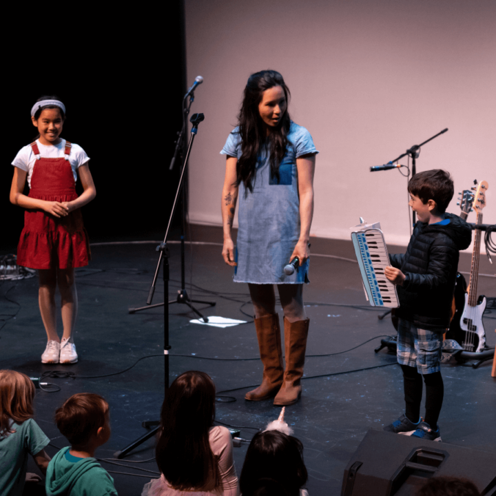 Ginalina on stage with a child in a black jacket holding an accordion and standing next to them, with another girl standing to the side