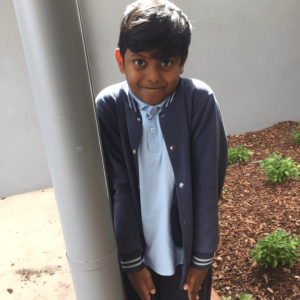 a boy, wearing a blue shirt and a dark grey jacket, leaning against a grey pole and standing on dirt