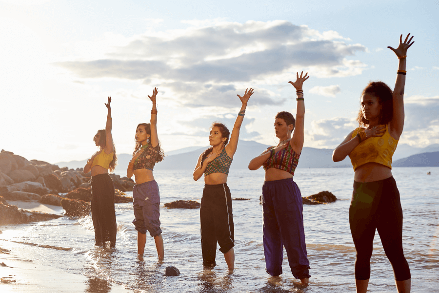 Her Tribal Roots dancers standing at the shore in the shallow water. With their left arm raised.