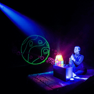 performer for the show ZOOOM, sitting in a dark room with a colorful glow in the dark shapes in the background
