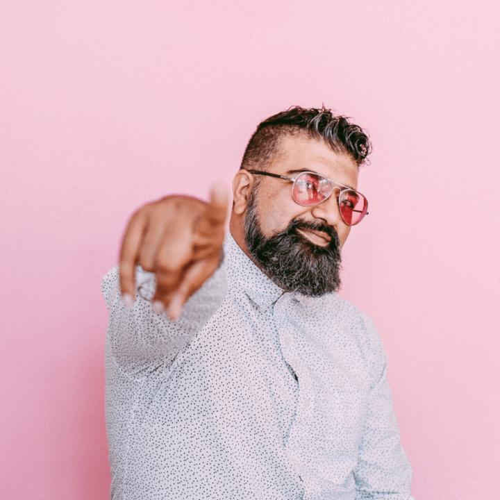 Ruploops pointing to the camera, while wearing a white shirt and pink tinted glasses, with a pink backdrop