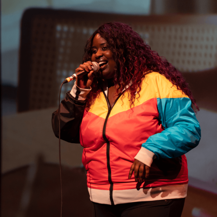 Missy D performing in stage, holding a microphone, and wearing a yellow, red, and blue jacket