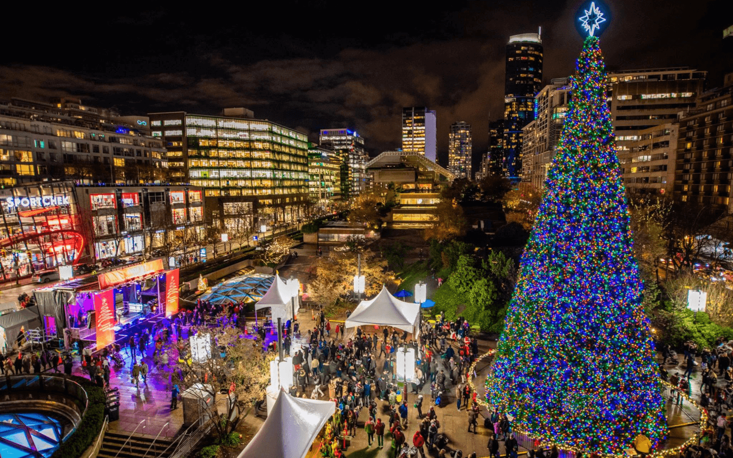 a large Christmas tree lit up at night, with crowds of people standing around it, with a buildings in the background.