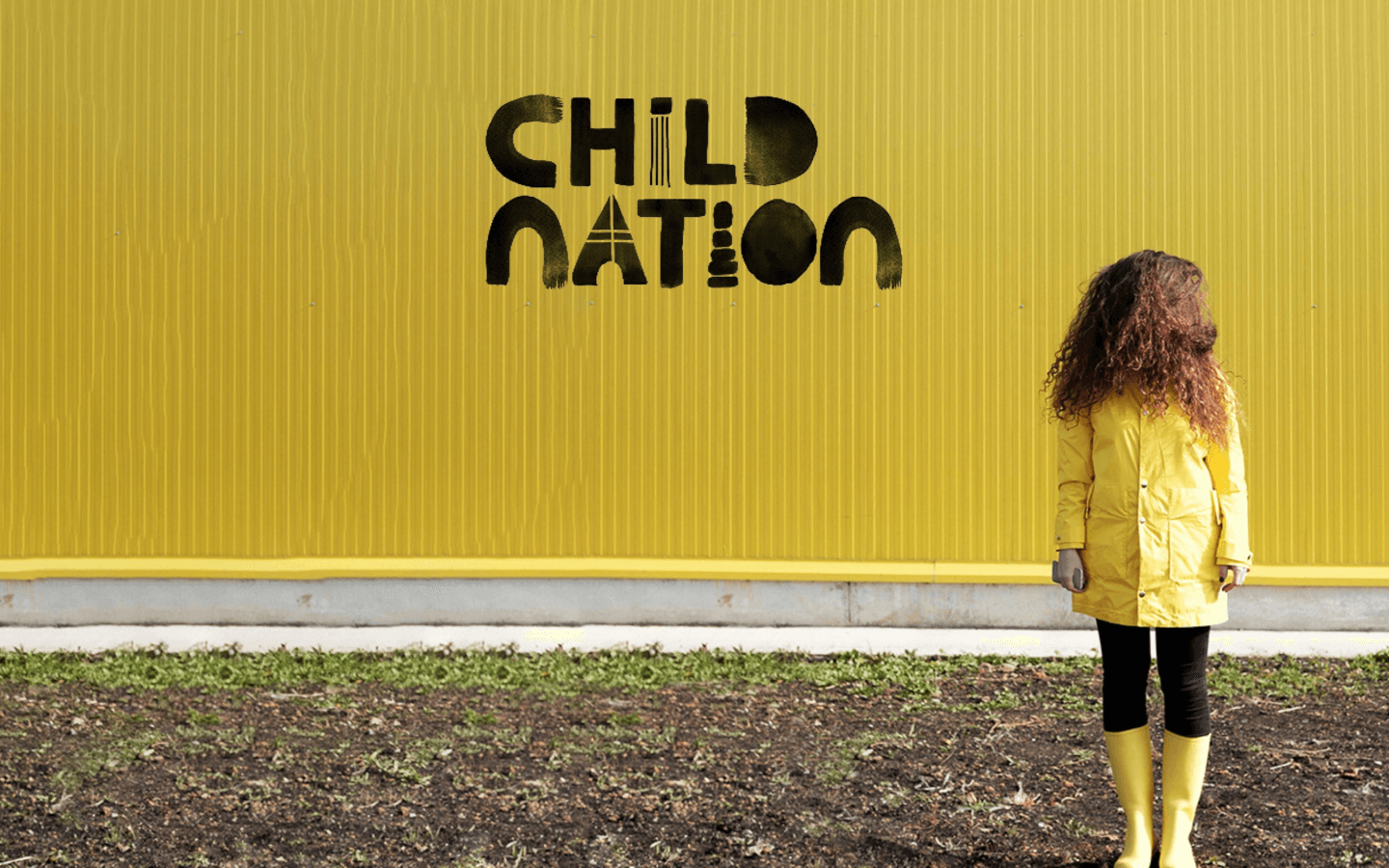 a child in a yellow rain coat and boots, standing in front of a yellow wall which has Child Nation written on it.