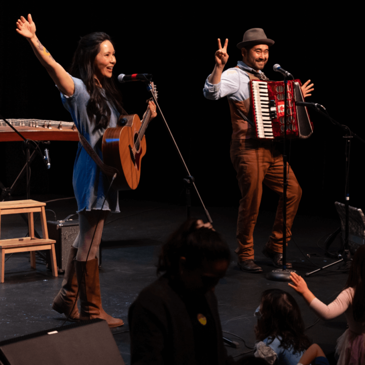 Ginalina on stage holding a guitar while singing, and other performer holding an accordion