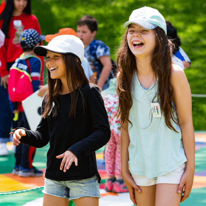 2 girls smiling while standing outside in the activity village, with school children in the background