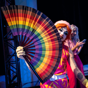 A performer from Glamily, holding a rainbow fan and smiling.